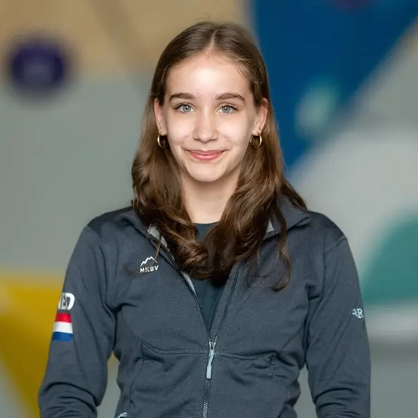 Een jonge vrouw met lang bruin haar, gekleed in een donker jasje met een patch in de vorm van de Nederlandse vlag, die glimlacht tegen een wazige, kleurrijke achtergrond.